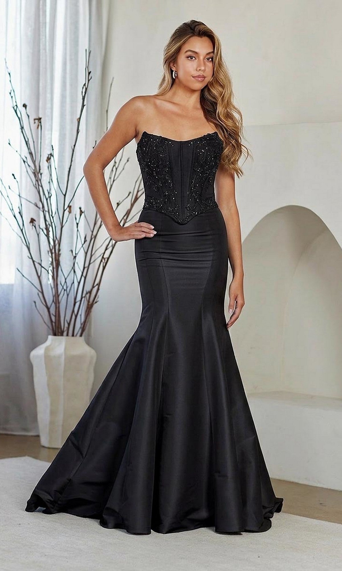 A woman poses indoors with her hand on her hip wearing the black Amelia Couture 8080 long prom dress, featuring a strapless embellished corset bodice and fit-and-flare skirt, against a neutral background with bare branches.